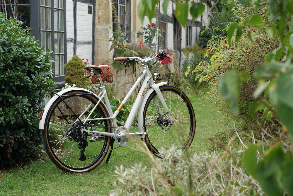 A cream-coloured classic e-bike with step through frame and brown leather saddle, parked on a green lawn in a cottage garden