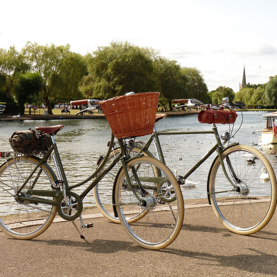 Two classic green bicycles, one with front wicker basket and one with leather bottle holder, parked by a riverside