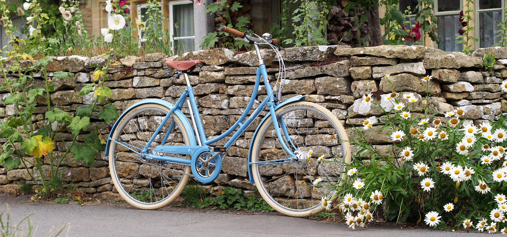 A blue Pashley Poppy leant against a dry stone wall with daisies and hollyhocks alongside.