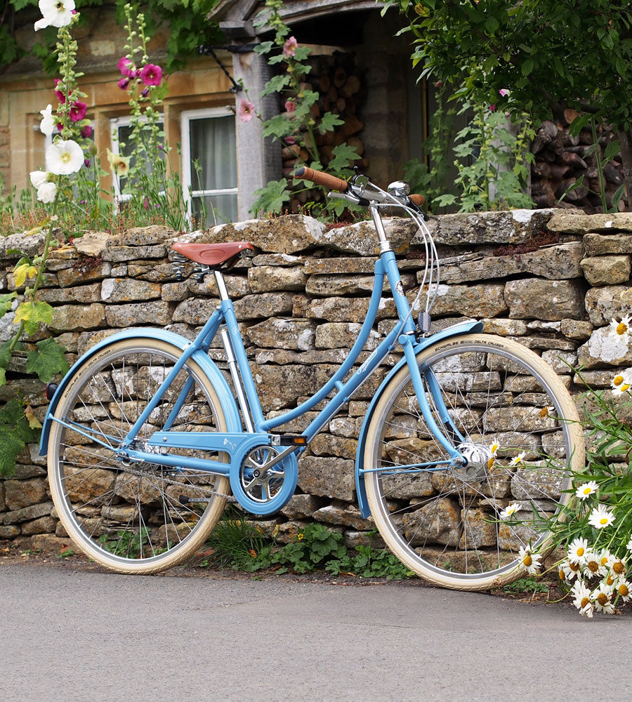 A blue Pashley Poppy leant against a dry stone wall with daisies and hollyhocks alongside.